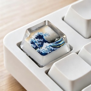 May include: A custom keyboard keycap featuring a design of Hokusai's "The Great Wave off Kanagawa." The keycap is clear with the artwork visible beneath a transparent top. The keycap is set in a white keyboard.