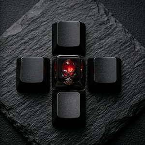 May include: A set of five black keycaps arranged in a cross shape. The central keycap is transparent, revealing a red gem and a skull design. The other four keycaps are solid black and square-shaped.