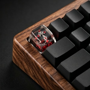 May include: Close-up of a custom keyboard keycap featuring a decorative cross design. The keycap is clear with a red and silver cross, set against a red background. The keyboard has a wooden frame and black keys.