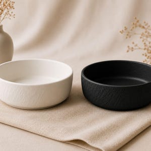 May include: Two ceramic bowls, one white and one black, sit side-by-side on a beige cloth. The bowls have a textured exterior and a smooth interior. The white bowl is on the left, and the black bowl is on the right.