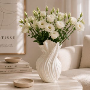 Op de afbeelding: Een witte keramische vaas met een uniek, wervelend ontwerp bevat een boeket witte bloemen. De bloemen hebben meerdere lagen bloemblaadjes en groene stelen. De vaas staat op een witte tafel met boeken en kleine kommen.