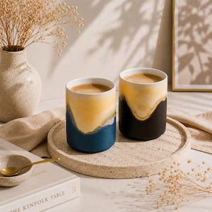 May include: Two cylindrical ceramic mugs filled with coffee. The left mug features a gradient of cream, soft pink, and deep blue. The right mug displays a gradient of cream and matte black. Both mugs have a smooth, rounded rim and base.