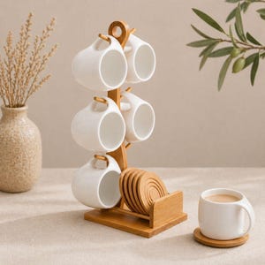 May include: A white ceramic mug set with a bamboo rack and coasters. The mugs have a unique handle design and are displayed on a natural wood rack. The set includes six mugs and coasters, perfect for coffee or tea.