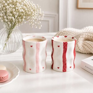 Pode incluir: Duas canecas de cerâmica com textura ondulada e nervurada. Uma caneca apresenta riscas verticais cor-de-rosa claro e pequenos corações vermelhos, enquanto a outra tem riscas verticais vermelho-escuras e pequenos corações vermelhos.