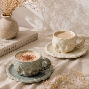 May include: Two ceramic cups and saucers with an irregular, dimpled texture. One cup is a muted blue-grey, and the other is a warm cream color. Both cups are filled with coffee and sit on matching textured saucers with scalloped edges.