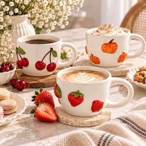 Può includere: Tre tazze in ceramica bianca con disegni di frutta in rilievo. Una tazza mostra ciliegie rosse, un'altra zucche arancioni con panna montata e la terza fragole rosse riempite con un latte macchiato con decorazione di schiuma.