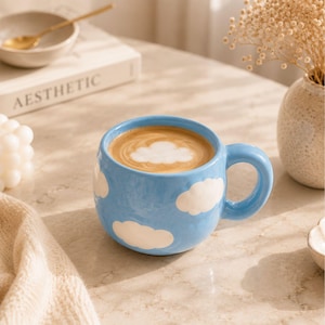 Pode incluir: Uma caneca de cerâmica redonda em azul claro com um padrão de nuvens brancas no exterior. A caneca está cheia de uma bebida de café com um design de espuma em forma de nuvem na superfície. Possui uma alça grossa e curva e acabamento brilhante.