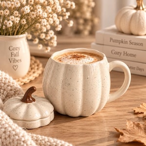 Peut inclure: Une tasse en céramique couleur crème en forme de citrouille avec une finition mouchetée et un couvercle assorti doté d'une tige marron. La tasse possède une anse incurvée et contient une boisson chaude avec de la mousse et de la cannelle.