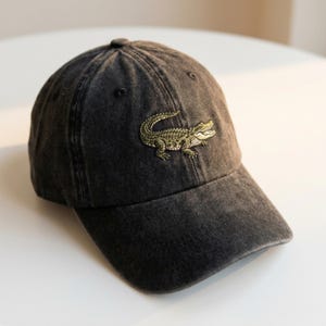 Könnte beinhalten: Eine schwarze Baseballkappe mit Used-Look. Auf der Vorderseite befindet sich ein grün-beiges, gesticktes Alligatorendesign. Die Kappe hat einen gebogenen Schirm und eine runde Krone.