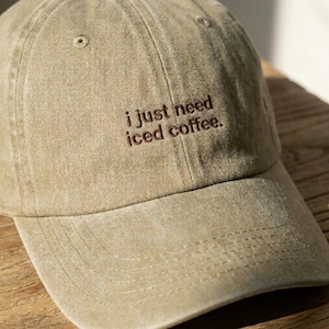 May include: A khaki baseball cap with the embroidered text "i just need iced coffee." The hat features a curved peak and a classic six-panel design. The hat is made of a soft, worn material.
