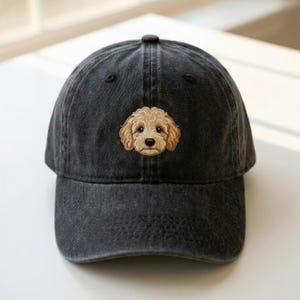 May include: A black baseball cap with a distressed finish. The cap features an embroidered image of a light brown dog's face on the front. The cap has a curved brim and an adjustable strap at the back.