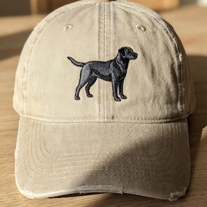 May include: A beige baseball cap with a distressed edge. It has an embroidered black Labrador Retriever dog on the front. The hat is made of a soft, worn material.