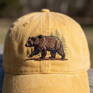 May include: Mustard yellow baseball cap featuring an embroidered brown bear walking to the left, with green pine trees behind it. The cap has a curved brim and a fabric strap closure. This cap is perfect for outdoor enthusiasts.