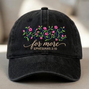 May include: Black baseball cap featuring embroidered text and floral embellishments. The text reads "MADE for more" in blue script, with pink flowers and green leaves. The scripture reference "Ephesians 2:10" is below the main text.