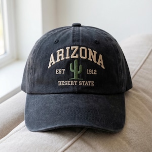 May include: A dark gray baseball cap with the word "ARIZONA" embroidered in off-white, along with a green cactus graphic. The text "EST 1912 DESERT STATE" is also embroidered on the cap. The cap is sitting on a beige surface.