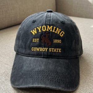 May include: A dark blue baseball cap with the word "WYOMING" embroidered in gold lettering. The cap also features the text "EST 1890 COWBOY STATE" and a brown image of a cowboy on a horse.