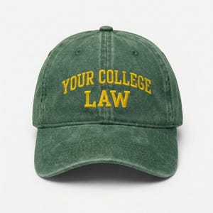 Peut inclure: Casquette de baseball verte avec l'inscription "YOUR COLLEGE LAW" brodée en jaune. La casquette a une visière incurvée et un aspect légèrement délavé. Le chapeau est en matière souple.
