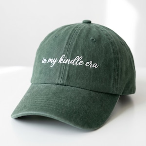 Peut inclure: Casquette de baseball vert foncé avec l'inscription brodée "in my kindle era" en écriture blanche. La casquette a une visière incurvée et un petit bouton sur le dessus. Le chapeau est en tissu doux et texturé.