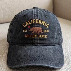 Op de afbeelding: Een zwarte baseball cap met goudkleurig geborduurd opschrift "CALIFORNIA", een bruin berenlogo en "GOLDEN STATE". De cap heeft ook de tekst "EST 1850".