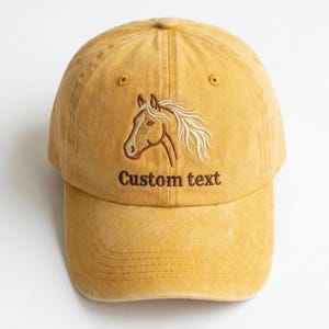 Peut inclure: Casquette de baseball jaune moutarde avec une tête de cheval brodée sur le devant. Le cheval est marron avec une crinière blanche. Le texte "Custom text" est brodé en dessous. Visière incurvée et sangle réglable.