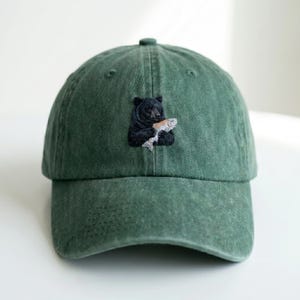 Peut inclure: Casquette de baseball verte avec un ours noir brodé tenant un poisson. La casquette a une visière incurvée et une fermeture à sangle en tissu. Le tissu de la casquette est doux et usé.