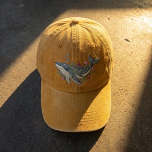 Op de afbeelding: Mosterdkleurige baseballcap met een geborduurd walvisontwerp. De walvis is afgebeeld in blauw- en groentinten, omringd door bloemaccenten in roze en paars. De cap heeft een gebogen klep en een klassiek zes-panelen ontwerp.