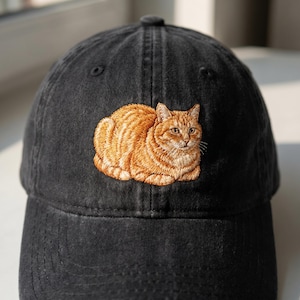 以下が含まれることがあります： オレンジ色のトラ猫のデザインが刺繍された黒い野球帽。猫はリラックスしたポーズで描かれており、毛並みや顔の表情が細かく表現されています。帽子は柔らかく、使い古された生地で作られています。
