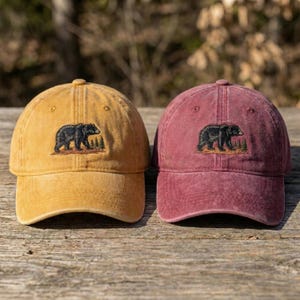 Puede incluir: Se muestran dos gorras de béisbol, una amarilla mostaza y otra burdeos. Cada gorra presenta un diseño bordado de un oso negro y un árbol. Las gorras están hechas de un material suave y desgastado, y están sobre una superficie de madera.