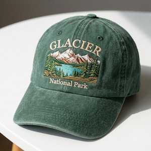 May include: A green baseball cap with an embroidered design featuring a mountain landscape, lake, and trees. The text "GLACIER National Park" is embroidered above the landscape. The cap has a curved brim and a button on top.