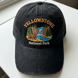May include: Black baseball cap with embroidered "Yellowstone National Park" design. The design features a bear, waterfall, and trees. The text is in yellow and white, with a blue river and green trees.