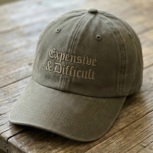 Puede incluir: Gorra de béisbol verde oliva con el texto bordado "Expensive & Difficult" en fuente gótica. La gorra tiene una visera curva y un cierre de correa de tela. Hecha de un material suave y duradero.