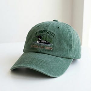 Op de afbeelding: Groene baseballpet met geborduurd ontwerp. Het ontwerp bevat het woord "MINNESOTA", een ijsvogel en de zin "L'ETOILE DU NORD". De pet is gemaakt van een zachte stof en heeft een gebogen klep.