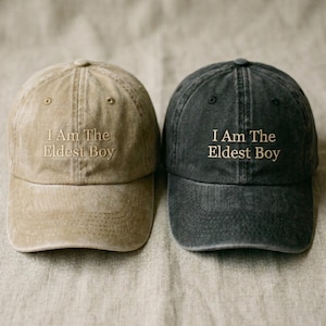 Op de afbeelding: Twee baseballpetten, één beige en één donkergrijs, worden naast elkaar getoond. Beide petten hebben de geborduurde tekst "I Am The Eldest Boy" in een bijpassende kleur. De petten zijn gemaakt van een zachte stof en hebben een klassiek zes-panelen ontwerp.