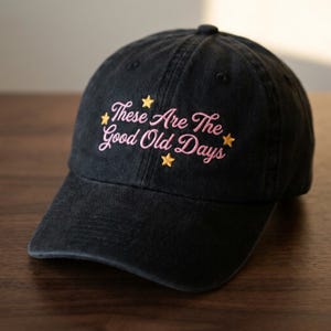 May include: Black baseball cap with the embroidered text "These Are The Good Old Days" in pink script and yellow stars. The cap is made of a soft, worn fabric and has a curved brim. A casual accessory.