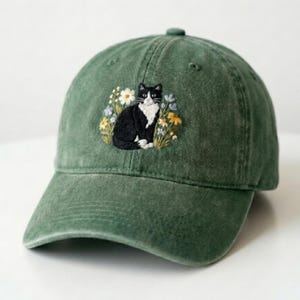 Op de afbeelding: Groene baseballcap met een geborduurd ontwerp van een zwart-witte kat omringd door gele, witte en blauwe bloemen. De cap heeft een gebogen klep en een stoffen bandsluiting.
