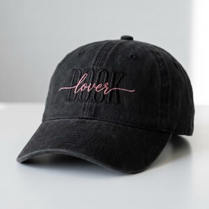 Peut inclure: Casquette de baseball noire avec le texte brodé "BOOK lover" en script rose. Le chapeau est fait d'un tissu doux et usé et a une visière incurvée. Un accessoire décontracté pour les amateurs de livres.