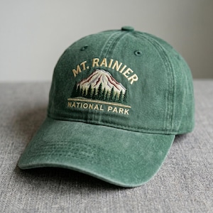 Può includere: Cappellino da baseball verde con la scritta "MT. RAINIER NATIONAL PARK" ricamata sul davanti. Il design presenta una montagna e alberi. Il cappellino è realizzato in un tessuto morbido e strutturato.