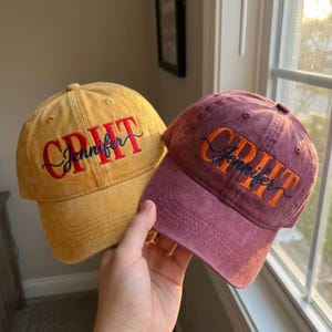 May include: Two baseball caps, one mustard yellow and one maroon, both featuring embroidered text. The yellow cap has red block letters spelling GRIT with the name Jennifer in navy blue script. The maroon cap has orange block letters spelling GRIT with the name Jennifer in navy blue script.