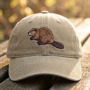Può includere: Cappellino da baseball beige con un castoro marrone ricamato. Il castoro ha una pelliccia marrone dettagliata e una coda marrone scuro. Il cappellino ha una visiera curva e un cinturino regolabile.