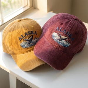 May include: Two baseball caps, one mustard yellow and one burgundy, are displayed. Both caps feature the word "ALASKA" embroidered in blue, along with a whale and ocean scene. The caps have a worn, vintage look.
