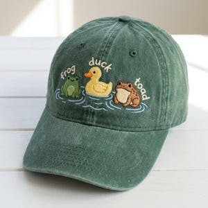 Könnte beinhalten: Grüne Baseballkappe mit gestickten Cartoon-Figuren: ein Frosch, eine Ente und eine Kröte. Die Wörter "frog", "duck" und "toad" sind über jeder Figur gestickt. Die Kappe hat einen gebogenen Schirm und einen Stoffriemen.