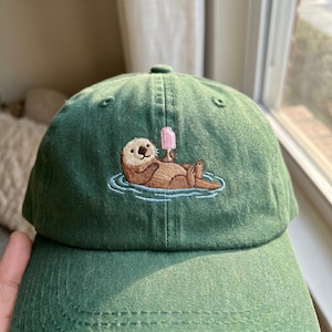 May include: Green baseball cap featuring an embroidered design of a brown otter relaxing in water and holding a pink ice lolly. The cap is made of a soft fabric and has a curved peak.