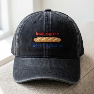 May include: A black baseball cap with the embroidered text "less regrets" in red, a baguette, and "more baguettes" in blue. The cap has a curved brim and a button on top. The fabric appears to be a worn, washed cotton.