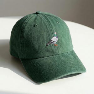 Puede incluir: Gorra de béisbol verde oscuro con visera curva. Presenta un bordado de una paloma en la parte delantera. La paloma tiene detalles grises, blancos, morados y naranjas. La gorra está hecha de una tela suave y texturizada.