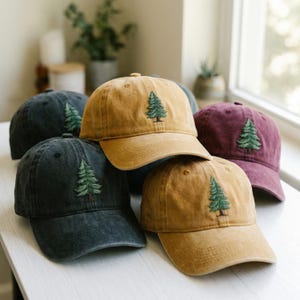 May include: A collection of baseball caps in various colors, including mustard yellow, dark gray, and burgundy. Each cap features an embroidered green pine tree design on the front. The caps are arranged on a white surface.
