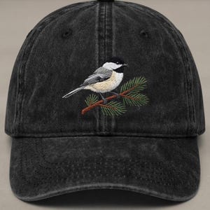 Op de afbeelding: Een zwarte, verwassen baseballcap met een geborduurd vogelontwerp. De vogel heeft zwarte, grijze en crèmekleurige veren, zittend op een bruine tak met groene naalden. De cap heeft een gebogen klep en een verstelbare band.