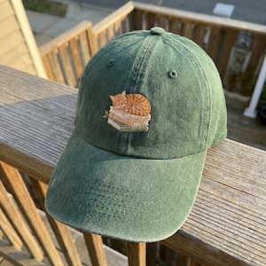 May include: A green baseball cap with a curved brim. The cap features an embroidered design of a sleeping orange tabby cat on a stack of books. The cap is made of a soft, textured fabric.