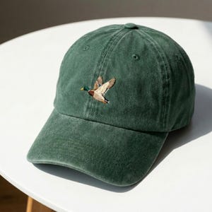 Könnte beinhalten: Dunkelgrüne Baseballkappe mit gebogenem Schirm. Die Kappe hat ein gesticktes Bild einer fliegenden Ente in Braun-, Beige- und Grüntönen. Die Kappe ist aus einem weichen, strukturierten Stoff gefertigt und hat einen klassischen, lässigen Stil.