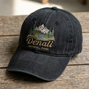 May include: Black baseball cap with an embroidered design featuring a mountain landscape, trees, and a lake. The text "Denali NATIONAL PARK" is embroidered in gold thread. The cap has a curved brim and a button on top.