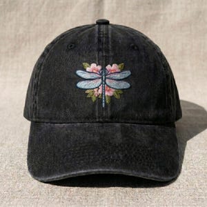 Puede incluir: Gorra de béisbol negra con un acabado desgastado. La gorra presenta un diseño de libélula bordada con alas azul claro, flores rosas y hojas verdes. Tiene una visera curva y una correa ajustable.